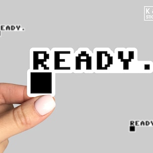 May include: A black and white sticker with the word "READY." in pixelated text. The sticker is on a gray background.