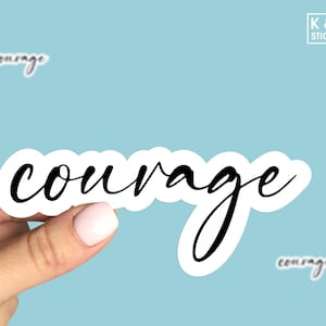 May include: White sticker with the word "courage" written in black cursive script. The sticker is being held by a hand.