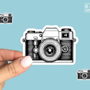 May include: Black and white illustration of a vintage camera with a lens and a flash. The camera is on a white background.