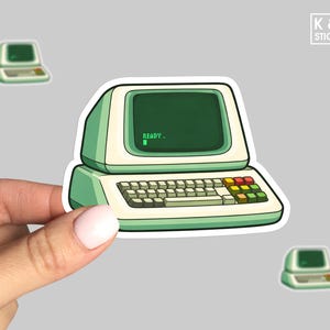 May include: A sticker featuring a retro computer with a green screen displaying "READY." The sticker has a white and green color scheme, with a keyboard and colorful function keys. The sticker is held by a hand.