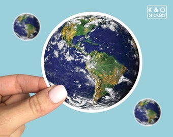 May include: A sticker of a blue and green Earth with clouds. The sticker is being held by a hand. The sticker is round and has a white border.