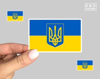 May include: A rectangular sticker featuring the blue and yellow flag of Ukraine with the yellow Tryzub coat of arms centered in the middle. The sticker has a white border and is displayed against a plain gray background with K&O STICKERS text.