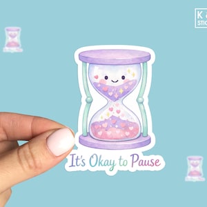 May include: A sticker featuring a watercolor hourglass with a smiling face and hearts. The hourglass is purple and pink, with the text "It's Okay to Pause" below. The sticker is held by a hand against a light blue background.