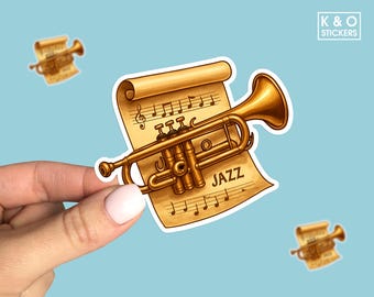 May include: A sticker featuring a golden trumpet and sheet music with the word "JAZZ". The sticker is held by a hand against a light blue background. Additional trumpet stickers are in the background. The sticker has a white border and the text "K & O STICKERS".