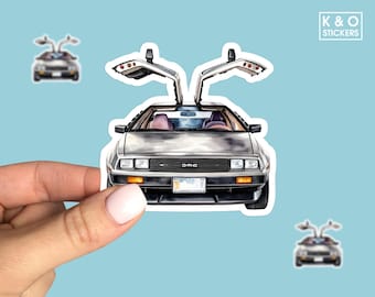 May include: A sticker featuring a silver DeLorean car with gull-wing doors open. The car has a black front bumper and the word "DMC" is visible. The sticker is held by a hand against a light blue background. The sticker has a white border.