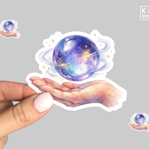May include: A sticker featuring a watercolor illustration of a hand cradling a purple and blue celestial orb with swirling rings. The sticker has a white border and the text "K & O STICKERS".