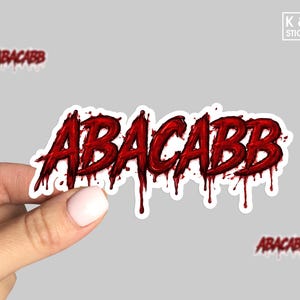 May include: A sticker with the word "ABACABB" in dripping red letters. The sticker is held by a hand against a gray background. The K & O Stickers logo is in the upper right corner.