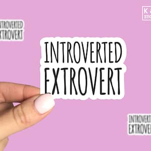 May include: White sticker with black text that reads "Introverted Extrovert". The sticker is being held by a hand.