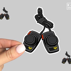 May include: A sticker featuring two black retro-style game controllers with red accents and a coiled black cable. Each controller has a circular dial and a small tennis racket graphic. The sticker is held by a hand.