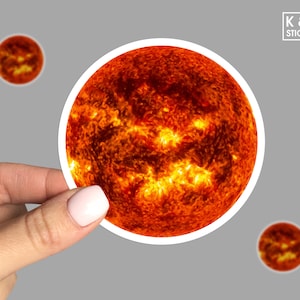 May include: A close-up of a sticker featuring a fiery orange and yellow image of the sun. The sticker is round and has a white border.
