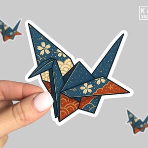 May include: A navy blue origami crane sticker with white floral and gold wave patterns, held by a hand. The sticker has red and gold accents. Two smaller crane stickers are also visible. The text "K & O STICKERS" is in the upper right corner.