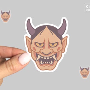 May include: A sticker featuring a tan-colored Japanese Oni mask with purple horns, sharp teeth, and a stern expression. The sticker is held by a hand. The image also includes two smaller versions of the same sticker and the text "K & O STICKERS".