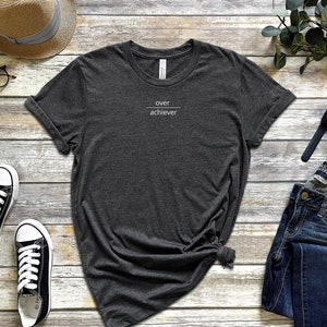 Op de afbeelding: Een donkergrijs T-shirt met de tekst "over achiever" erop gedrukt. Het shirt ligt op een houten oppervlak met een paar zwarte sneakers, een strohoed, een spijkerbroek, een horloge en een paar oordopjes.