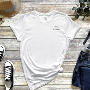 Op de afbeelding: Wit T-shirt met de tekst "over achiever" bedrukt op de voorkant. Het shirt ligt plat op een houten oppervlak met een paar zwarte sneakers, een strohoed, een bril, een blauwe jeans, een horloge en een paar oordopjes.