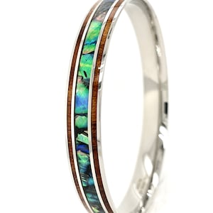 Puede incluir: Una pulsera de brazalete plateada pulida con una incrustación central de concha de abulón y madera oscura. La concha de abulón muestra iridiscentes azules, verdes y púrpuras. La madera es de color marrón oscuro y la pulsera tiene una superficie lisa y reflectante.
