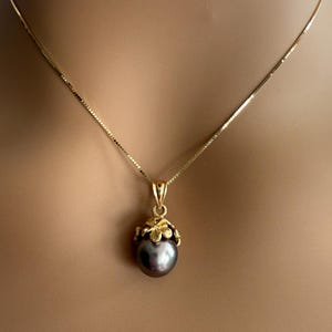 Puede incluir: Un collar de oro con un colgante de perla gris oscuro. El colgante est&aacute; engastado en un dise&ntilde;o floral dorado. La cadena es una fina cadena de oro. El collar se muestra en el cuello de una persona.