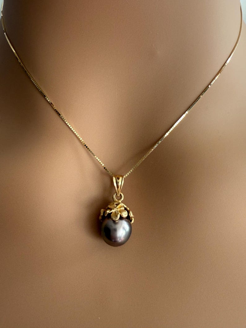 Puede incluir: Un collar de oro con un colgante de perla gris oscuro. El colgante est&aacute; engastado en un dise&ntilde;o floral dorado. La cadena es una fina cadena de oro. El collar se muestra en el cuello de una persona.