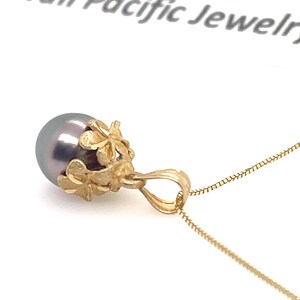 Puede incluir: Un collar con colgante de oro con una perla gris oscuro. La perla est&aacute; engastada en un dise&ntilde;o floral dorado. La cadena del collar es de color dorado delicado. Las palabras "Hawaii Pacific Jewelry" son visibles en el fondo.
