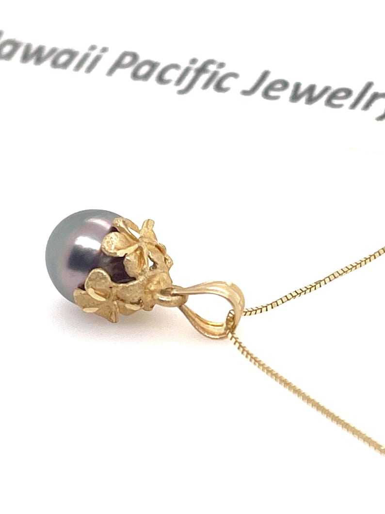 Puede incluir: Un collar con colgante de oro con una perla gris oscuro. La perla est&aacute; engastada en un dise&ntilde;o floral dorado. La cadena del collar es de color dorado delicado. Las palabras "Hawaii Pacific Jewelry" son visibles en el fondo.