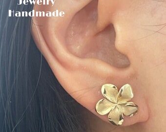May include: A close-up of a person's ear with a gold-colored flower-shaped stud earring. The earring is a detailed, five-petal design. The text "Hawaii Pacific Jewelry Handmade" is visible in the image.
