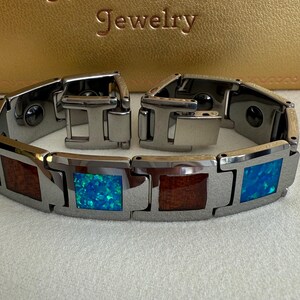 May include: A stainless steel bracelet with alternating rectangular inlays of wood and blue opal. The bracelet is open, showing the clasp mechanism. The background features the text "Hawaii Pacific Jewelry" in gold lettering.