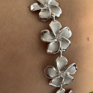 May include: A silver bracelet featuring a series of flower-shaped links. Each flower has five petals and a textured surface, creating a delicate and elegant design. The bracelet is shown on a person's wrist.