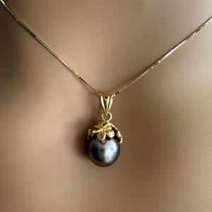 Puede incluir: Un collar dorado con un colgante de perla gris oscuro. El colgante est&aacute; engastado en un dise&ntilde;o floral dorado. La cadena es una fina cadena de caja dorada. El collar se muestra sobre un fondo neutro.