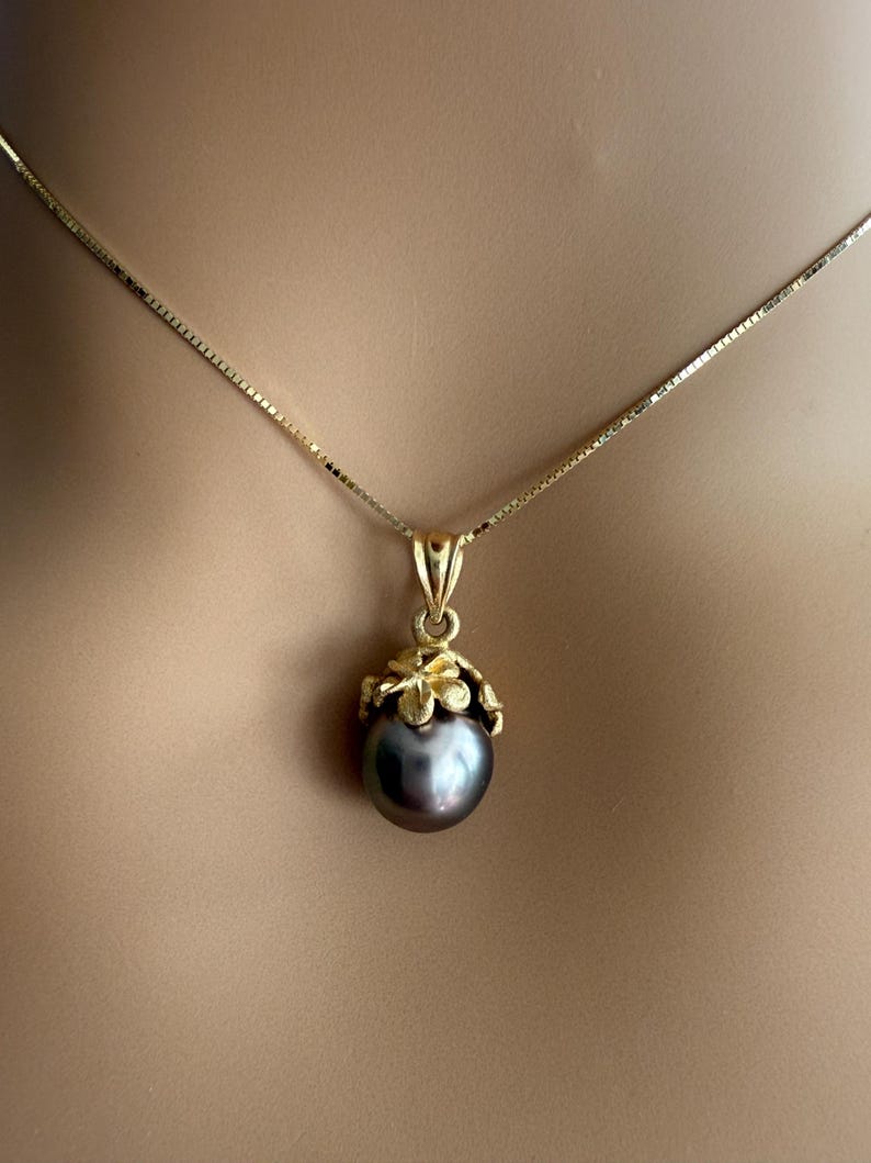 Puede incluir: Un collar dorado con un colgante de perla gris oscuro. El colgante est&aacute; engastado en un dise&ntilde;o floral dorado. La cadena es una fina cadena de caja dorada. El collar se muestra sobre un fondo neutro.
