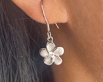 Può includere: Un orecchino a forma di fiore argentato con una piccola gemma trasparente al centro. L'orecchino è a goccia, con un gancio per orecchie forate. Il fiore ha cinque petali e una superficie strutturata.