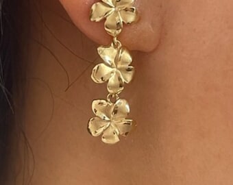 May include: Gold-toned floral drop earrings. The earrings feature three flower-shaped charms, each with detailed petal designs. The earrings are shown worn on an ear. The text "Hawaii Pacific Jewelry" is at the bottom of the image.