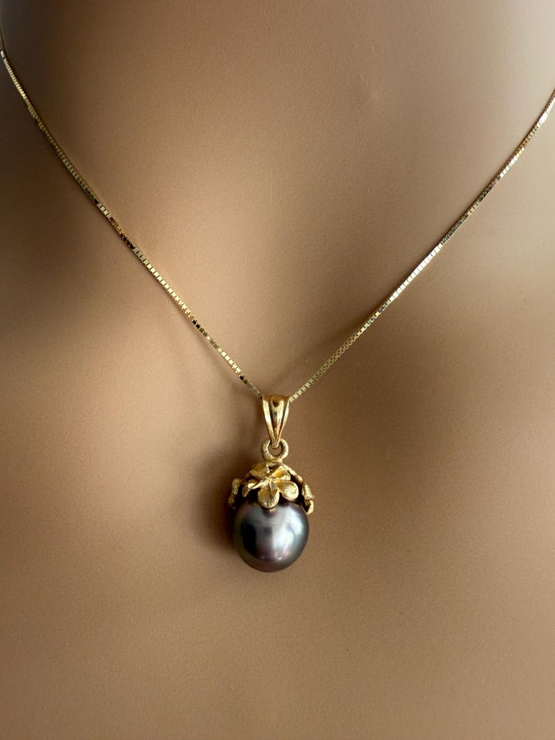 Puede incluir: Un collar de oro con un colgante con una perla gris oscuro. La perla est&aacute; encerrada en un dise&ntilde;o floral dorado, y la cadena es una delicada cadena de oro. El collar se muestra sobre un fondo neutro.