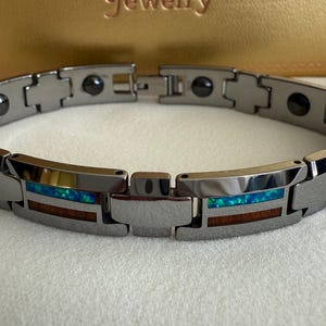 以下が含まれることがあります： 長方形のリンクが付いたシルバーカラーのブレスレット。各リンクには、青と緑のオパールのような素材と、茶色の木のような素材のセクションがあります。ブレスレットは白い表面に表示されています。