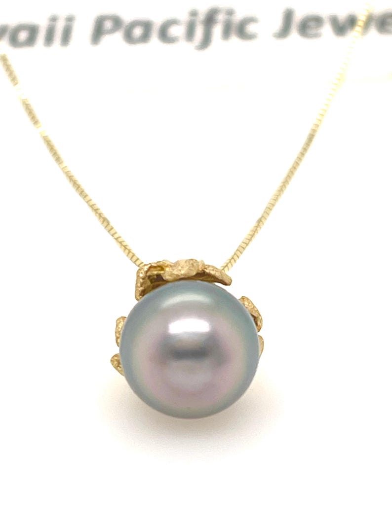 Puede incluir: Un collar de oro con un gran colgante de perla iridiscente. La perla tiene un suave tono gris-azul con un toque de rosa. El colgante est&aacute; unido a una delicada cadena de oro. El collar se muestra sobre un fondo blanco.