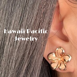 May include: Gold-tone flower-shaped earrings. The earrings are in the shape of a plumeria flower. The earrings are worn by a person with long dark hair. The text "Hawaii Pacific Jewelry" is visible in the image.
