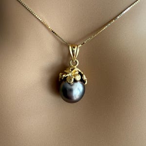 Puede incluir: Un collar dorado con un colgante de perla gris oscuro. El colgante est&aacute; adornado con un dise&ntilde;o floral dorado. La cadena es una fina cadena de caja dorada.
