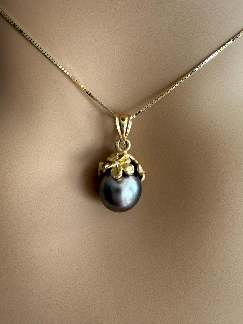 Puede incluir: Un collar dorado con un colgante de perla gris oscuro. El colgante est&aacute; adornado con un dise&ntilde;o floral dorado. La cadena es una fina cadena de caja dorada.