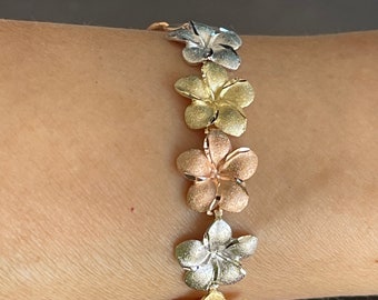 May include: A gold, silver, and rose gold bracelet with a chain of plumeria flower charms.