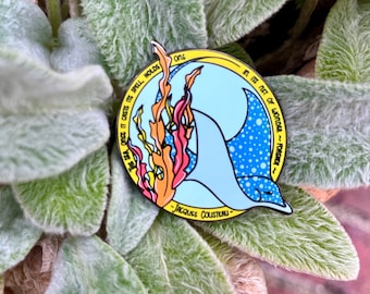 May include: A blue and white enamel pin with a manta ray and a quote by Jacques Cousteau: "The sea once it casts its spell holds its net of wonder - forever".