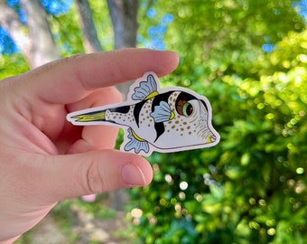 May include: A white, black, and yellow pufferfish sticker with blue fins. The sticker has a cartoon-like style and is held in a hand against a blurred background of green leaves.