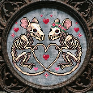 Könnte beinhalten: Ein gerahmtes Kreuzstichbild mit zwei Skelettmäusen, die ein Herz bilden. Die Mäuse sind beige und weiß, mit rosa Akzenten. Der Rahmen ist dunkelbraun mit einem verzierten Design. Der Hintergrund ist grau, mit roten Herzen und rosa Kreuzstichblumen.
