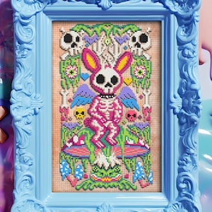 Può includere: Un'opera d'arte incorniciata con un coniglietto scheletro rosa con testa di teschio, circondato da teschi, funghi e fogliame. L'opera è in una cornice azzurra ornata. Lo sfondo include un teschio, una croce e altri elementi decorativi.