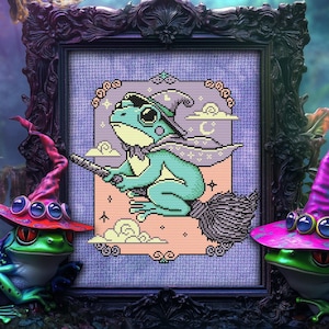 May include: A green frog wearing a witch's hat and riding a broomstick is depicted in a cross-stitch pattern framed in a dark wood frame. Two green frogs wearing witch hats are on either side of the frame.