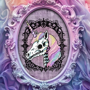 May include: A framed cross-stitch embroidery of a pastel-colored unicorn skull with a black lace design. The unicorn's horn is gold and the background is a light pink.