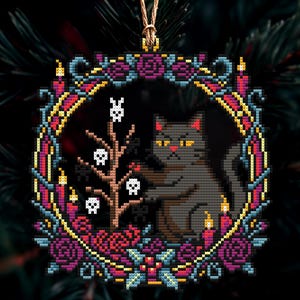 Op de afbeelding: Een kruissteek kerstversiering met een grijze kat met gele ogen, omringd door een krans van paarse rozen, kaarsen en een boom met schedels. De ornament is vierkant en heeft een gouden ophangkoord.