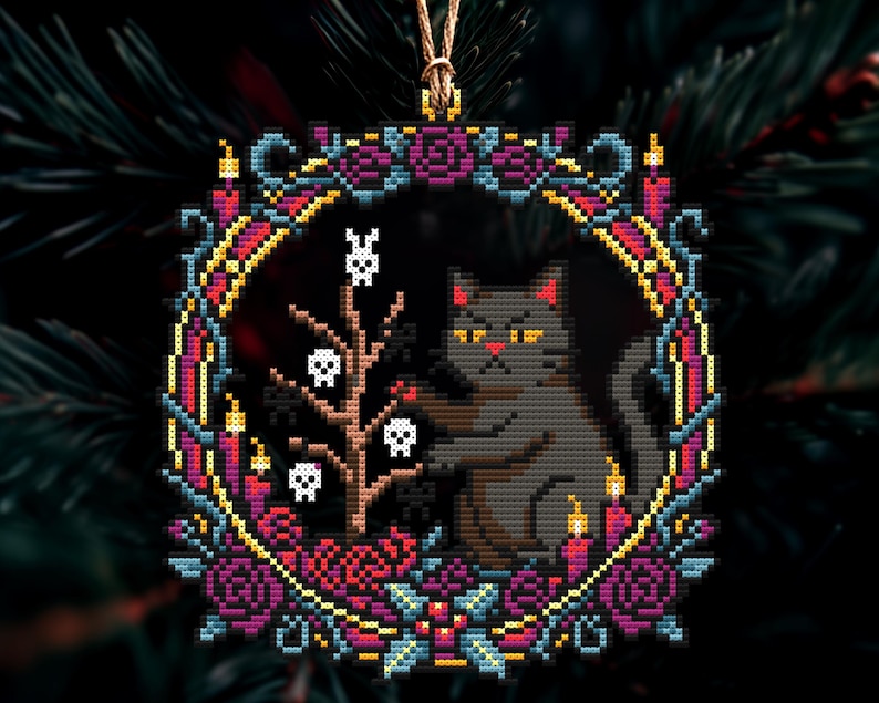 Op de afbeelding: Een kruissteek kerstversiering met een grijze kat met gele ogen, omringd door een krans van paarse rozen, kaarsen en een boom met schedels. De ornament is vierkant en heeft een gouden ophangkoord.