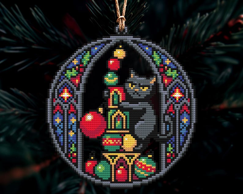Op de afbeelding: Een kruissteek kerstversiering met een zwarte kat met gele ogen, omringd door kleurrijke ballen. De versiering heeft een glas-in-lood stijl rand met rode, blauwe en groene accenten. Een gouden draad is bevestigd om op te hangen.