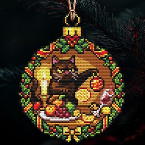 Op de afbeelding: Kruissteek kerstbal met een bruine kat die naar een rood-gouden bal grijpt. De kat is omringd door een tafel met eten, een kaars en een glas wijn. De bal is omlijst met een groene en rode krans.