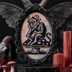 May include: A framed cross-stitch artwork featuring a black and white pixelated devil figure seated on a mushroom. The artwork is set in an ornate black oval frame. The background includes candles, a skull, and decorative wings, creating a gothic theme.