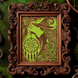 May include: A framed cross-stitch artwork featuring a witch silhouette holding a dreamcatcher. The artwork is in shades of green and burgundy, set within an ornate, dark brown wooden frame. The overall theme is mystical and witchy.