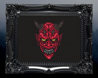 May include: A framed cross-stitch featuring a red devil face with horns, sharp teeth, and yellow eyes. The artwork is set against a black background and framed in a black ornate frame, evoking a gothic aesthetic.